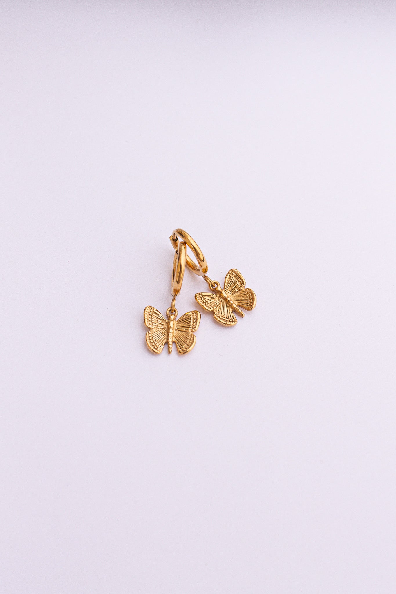A pair of gold-colored huggie hoop earrings with butterfly charms attached, displayed against a white background.